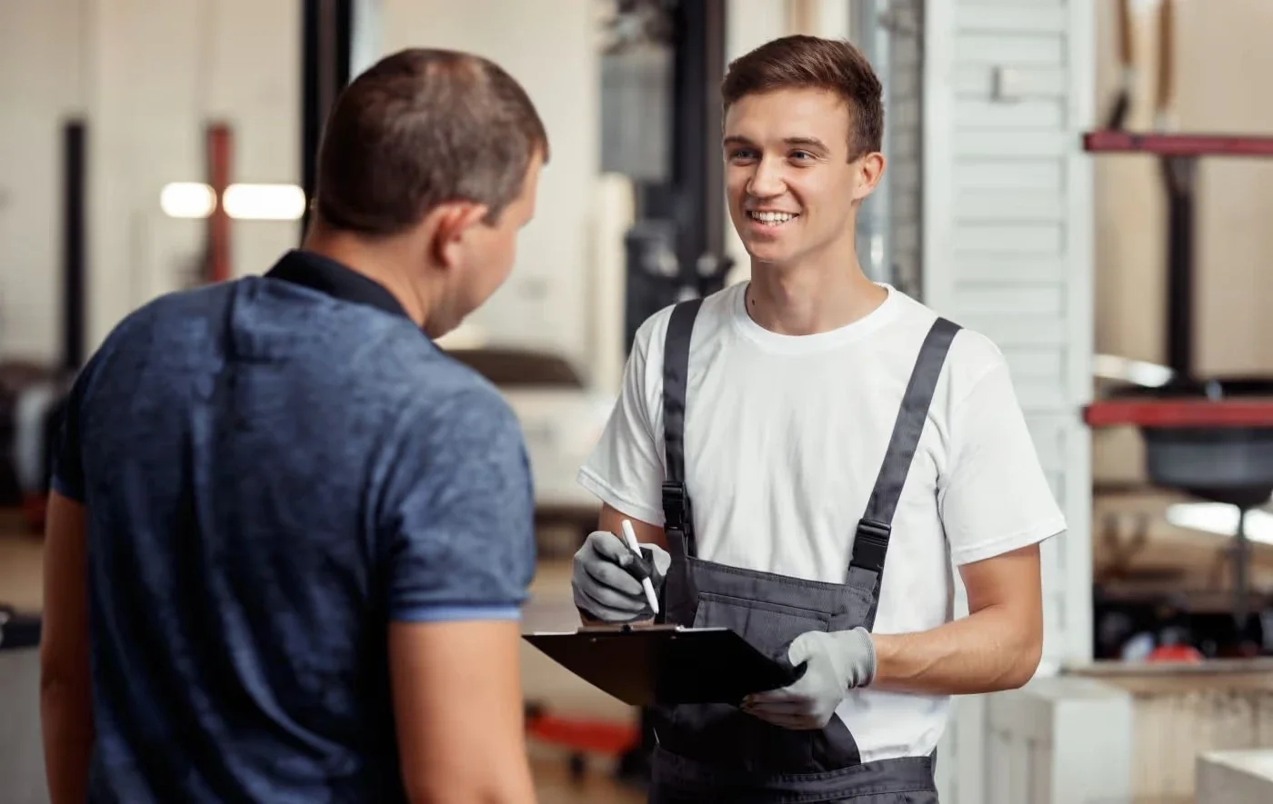 Smiling mechanic in overalls holding a clipboard, talking to a customer in a workshop.