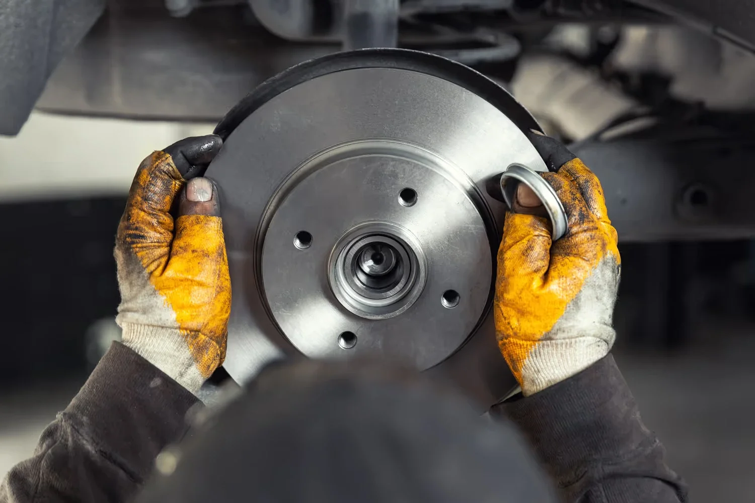 Person in dirty gloves installing a new brake rotor onto a car's wheel hub.