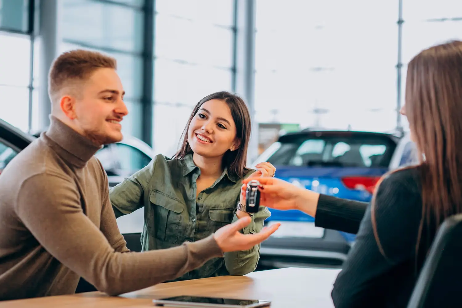 A couple smiling as a person hands them car keys in a dealership, signifying a purchase.