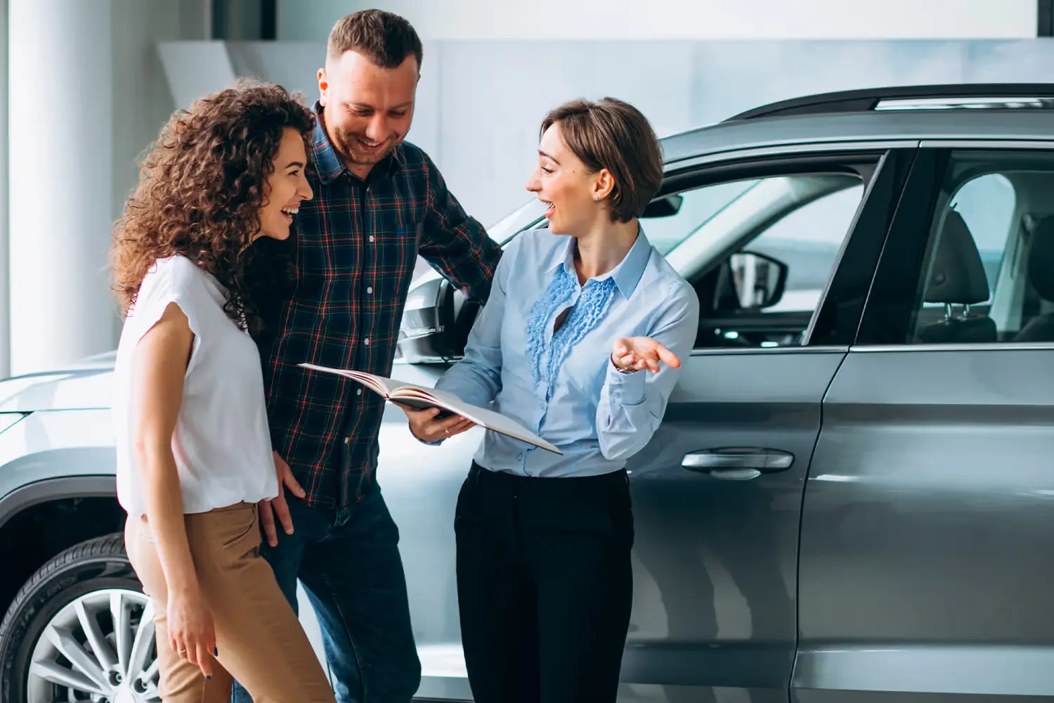 Car salesperson shows a brochure to a smiling couple next to a new grey SUV in a showroom.
