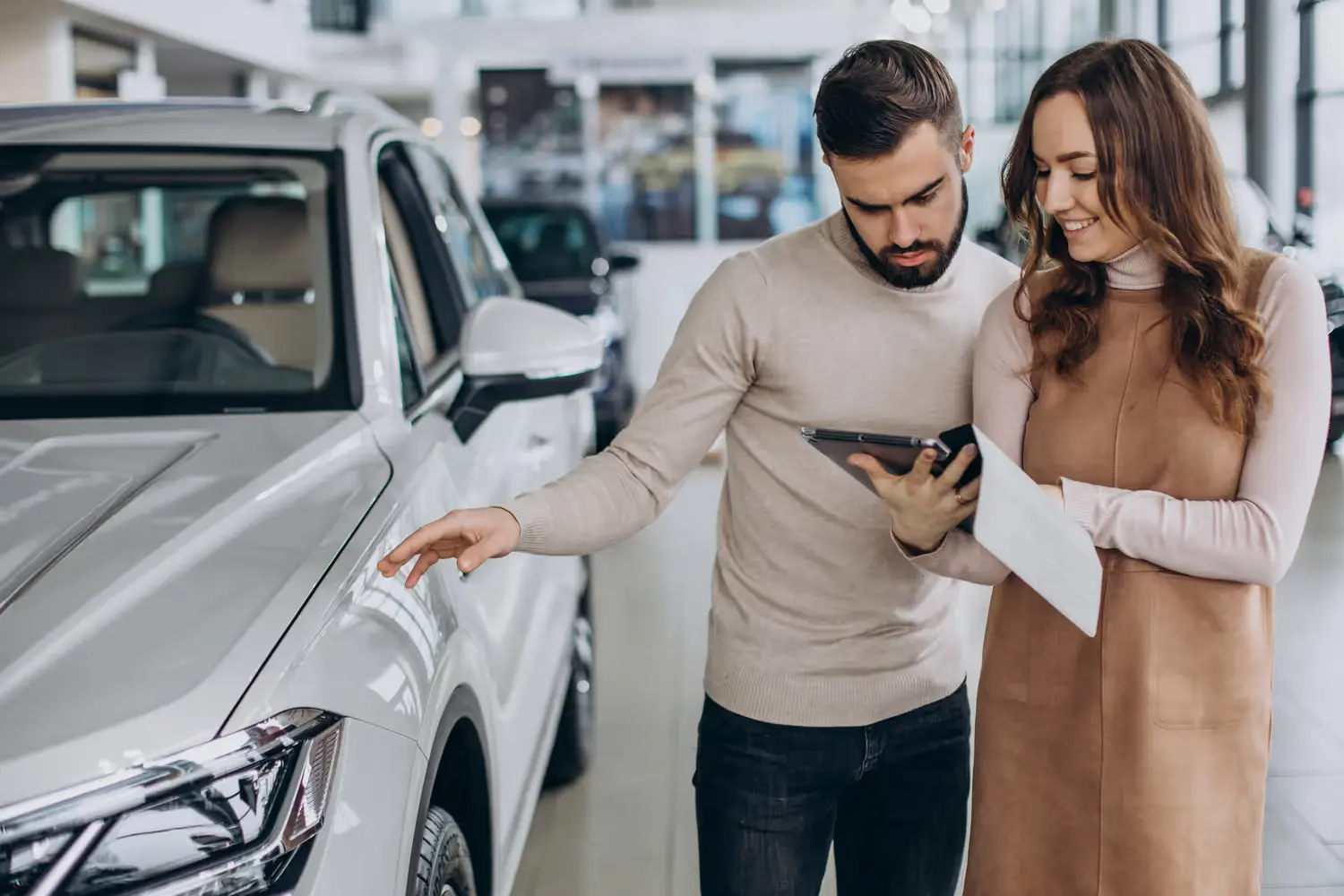 A man and woman looking at a white car in a showroom, discussing details from a tablet and papers.
