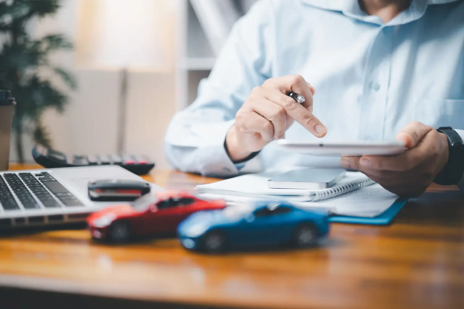 Person using a tablet at a desk with a laptop, car keys, and red and blue toy cars, suggesting vehicle insurance or sales.