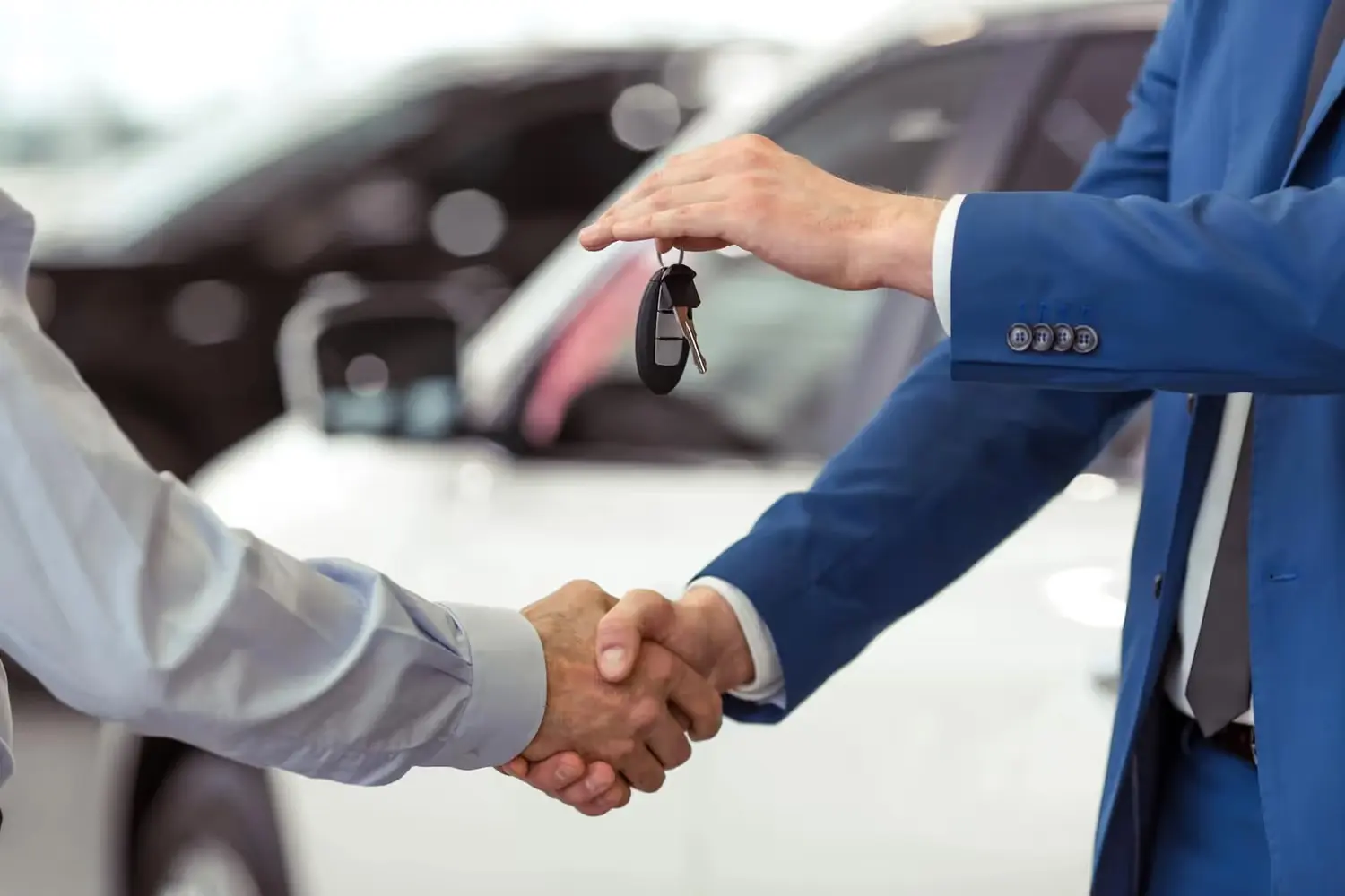 Two men shaking hands, one holding car keys, with cars blurred in the background.