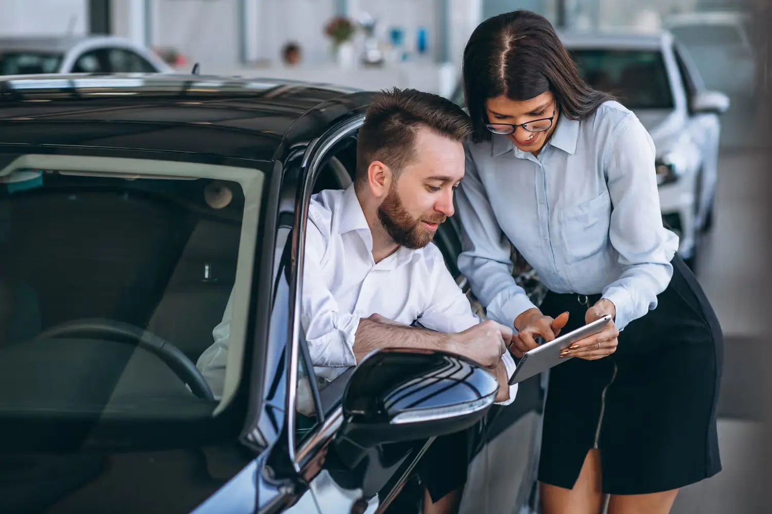 Man in car looking at tablet held by woman, likely at a car dealership.
