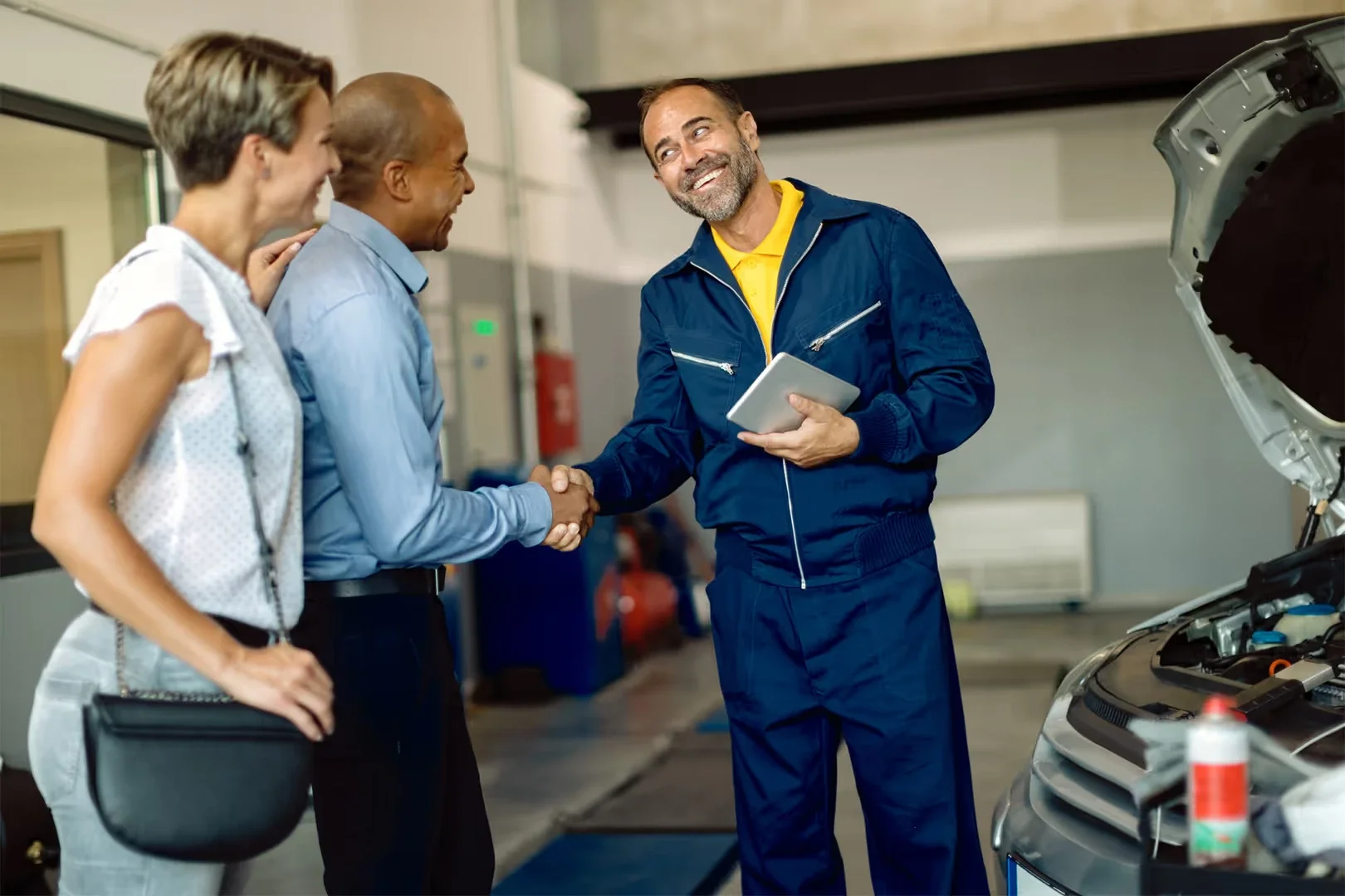 A smiling mechanic with a tablet shakes hands with a customer in a garage, a woman stands beside the customer.