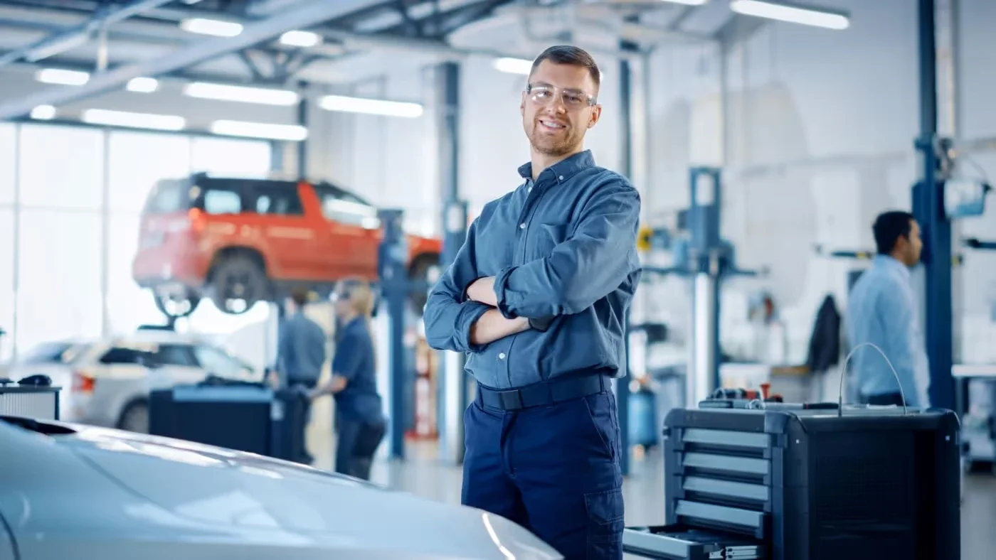 Smiling male mechanic in safety glasses and blue uniform, arms crossed, standing in a busy car repair shop.