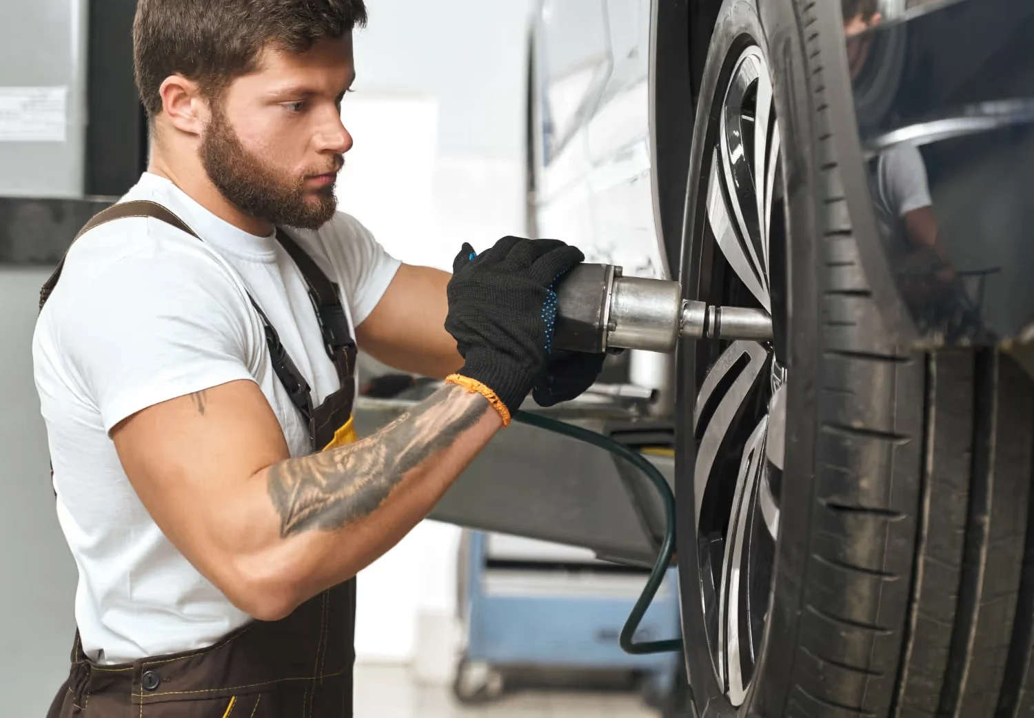 Mechanic in overalls and gloves using an impact wrench on a car wheel, which is elevated in a garage.